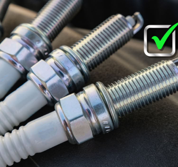 Four spark plugs lined up on a dark surface with a green checkmark symbol above them.